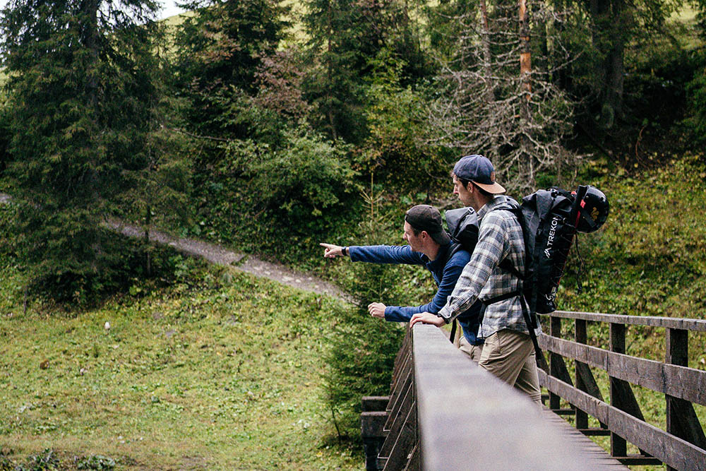 Two friends hiking along the Lech Trail in Austria, standing on a wooden bridge overlooking the river and enjoying the stunning alpine views